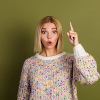 Young woman in a colorful knit sweater showing surprise while pointing up, set against a khaki green background.